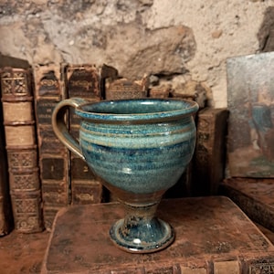 Peut inclure: Une tasse en céramique avec une base sur pied et une anse unique, présentant un vernis brillant dans des tons bleu profond, sarcelle et brun terreux. La tasse a une forme de bol arrondi avec des stries horizontales et un bord évasé.