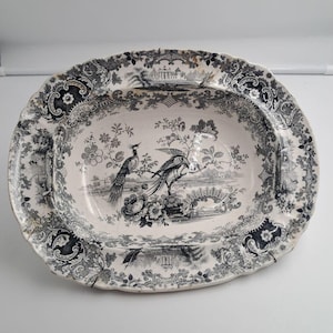 Peut inclure: Un plat de service ovale en céramique avec un motif de transfert noir et blanc. Le design représente deux oiseaux perchés sur une branche parmi des éléments floraux, avec un paysage en arrière-plan et une bordure festonnée ornée de motifs.
