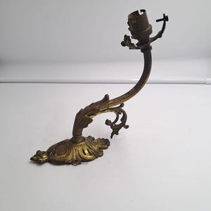 Puede incluir: Un aplique de pared de latón con un brazo curvo adornado con detalles en forma de hoja. La base es de forma ovalada con grabados decorativos, y la parte superior incluye un componente de casquillo metálico con un pequeño tornillo de ajuste.