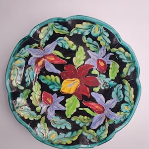 Könnte beinhalten: Ein runder Keramikt runder Keramikteller mit gewelltem Rand, der einen schwarzen Hintergrund mit handgemalten Blumenmotiven in Rot-, Violett- und Gelbtönen aufweist, umgeben von grünen Blättern mit goldfarbenen Umrandungen und einem blaugrünen Rand.