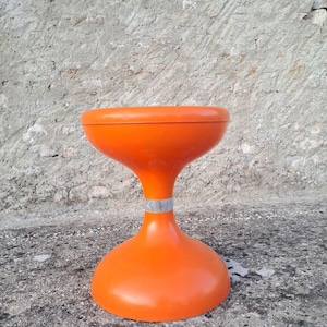 Op de afbeelding: Een feloranje kruk met een glad, zandlopervormig silhouet. De kruk heeft een brede, ronde zitting en een bijpassende brede voet, verbonden door een smal, taps toelopend middengedeelte met een dunne, metallic zilveren band in het midden.