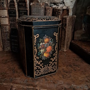 May include: A black hexagonal metal tin featuring a floral bouquet illustration in shades of orange, blue, and green. The container is decorated with gold-coloured filigree patterns along the edges and corners, and it includes a matching lid.