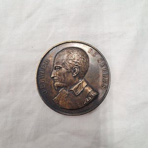 May include: A circular bronze-toned commemorative medal featuring a relief profile portrait of a man with a beard and period clothing. The text OLIVIER DE SERRES is embossed along the curved edge of the medal, which has a dark patina finish.
