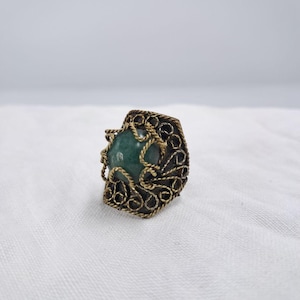 Peut inclure: Une bague ornée d'une pierre ronde vert foncé sertie dans une monture en métal sombre oxydé. Le sertissage est décoré d'un filigrane complexe en fil torsadé de couleur dorée qui entoure la pierre et recouvre les côtés de l'anneau.