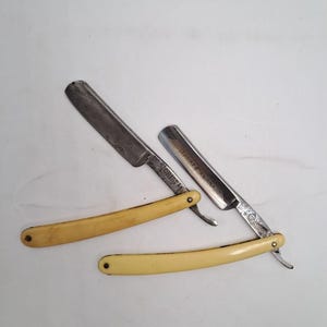 May include: Two vintage straight razors with cream-colored handles and metallic blades. One blade is engraved with the word CRISTA, while the other features the text ROYAL CANAILLE. Both razors have a folding design with visible metal rivets.