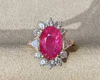 Può includere: Un anello in oro con una grande pietra preziosa rosa vibrante dal taglio ovale al centro. La pietra centrale è circondata da un alone di piccoli diamanti bianchi rotondi a taglio brillante, con un diamante bianco a taglio triangolare ai lati.