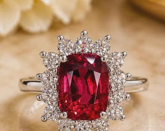 Può includere: Un anello con una grande pietra preziosa rosso intenso, di taglio rettangolare a cuscino al centro, circondata da un alone di dodici diamanti bianchi rotondi a taglio brillante su una fascia in metallo argentato lucido.