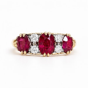 Könnte beinhalten: Ein Goldring mit drei oval geschliffenen roten Rubinen und vier rund geschliffenen weißen Diamanten. Der mittlere Rubin ist der größte, flankiert von zwei kleineren Rubinen an den Seiten, mit je zwei Diamanten zwischen den Rubinen in Krappenfassung.