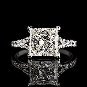 Peut inclure: Une bague argentée avec un grand diamant taille princesse carré au centre. L'anneau se divise en deux sections près du sertissage, les deux côtés étant incrustés de petites pierres rondes et transparentes le long de la tige de la bague.