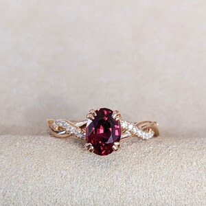 Pu&ograve; includere: Un anello in oro rosa con una pietra preziosa granato rosso scuro dal taglio ovale fissata da quattro griffe. La fascia presenta un design intrecciato con piccoli diamanti bianchi incastonati a pav&eacute; lungo i fili metallici che si intrecciano.