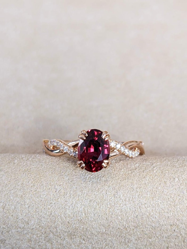 Pu&ograve; includere: Un anello in oro rosa con una pietra preziosa granato rosso scuro dal taglio ovale fissata da quattro griffe. La fascia presenta un design intrecciato con piccoli diamanti bianchi incastonati a pav&eacute; lungo i fili metallici che si intrecciano.