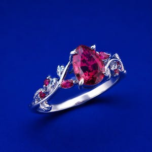 May include: A silver-toned ring featuring a large pear-cut deep red gemstone as the centerpiece. The band is designed with intricate metal leaf motifs and smaller marquise-cut red stones embedded along the sides, creating a botanical aesthetic.