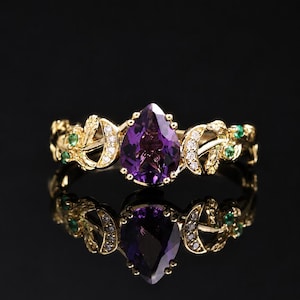 Op de afbeelding: Een gouden ring met een centrale peervormige paarse amethist vastgezet met klauwen. De band is ontworpen met halve maanvormen, kleine heldere steentjes en bladmotieven geaccentueerd met piepkleine groene steentjes voor een gedetailleerd uiterlijk.