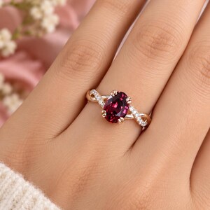 Pu&ograve; includere: Un anello in oro rosa con una pietra preziosa rosso scuro dal taglio ovale fissata da quattro griffe. La fascia presenta un design intrecciato con piccole pietre bianche incastonate a pav&eacute; lungo le sezioni che si incrociano, creando un aspetto dettagliato.