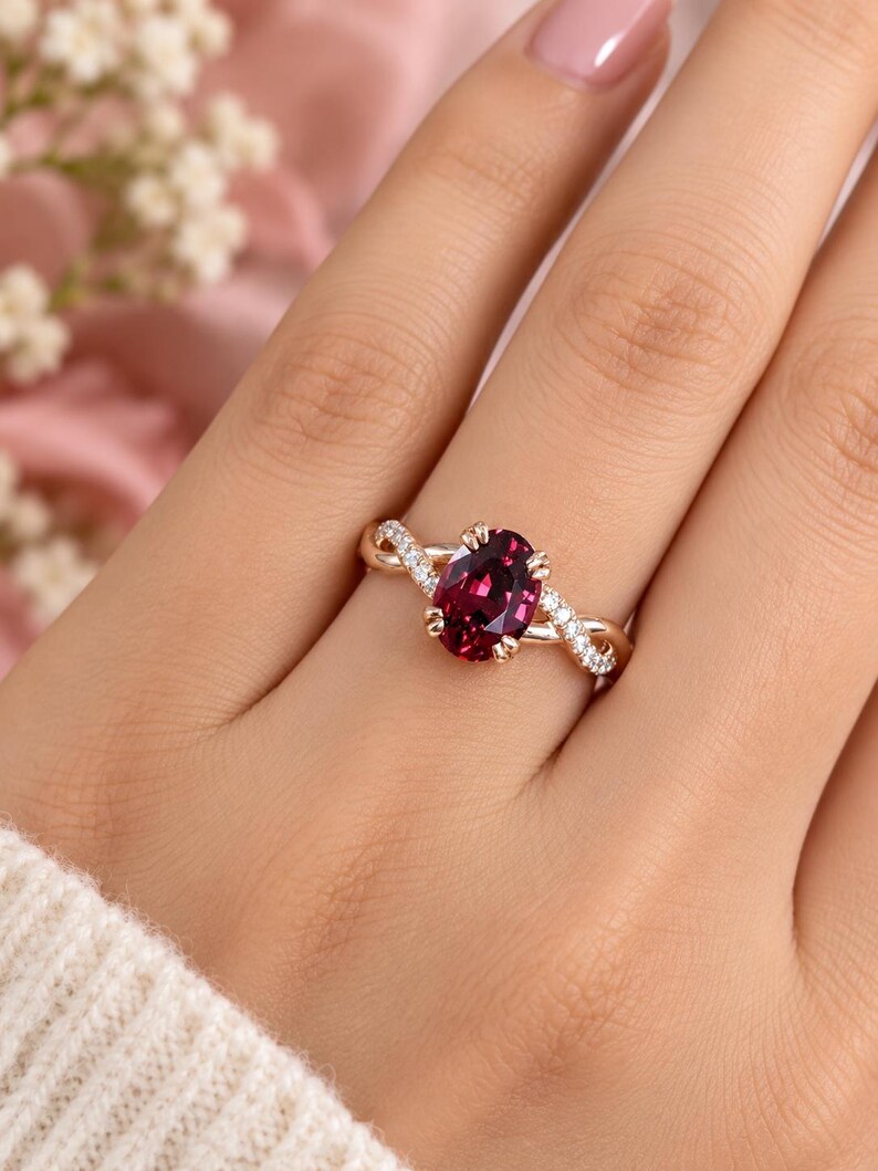 Pu&ograve; includere: Un anello in oro rosa con una pietra preziosa rosso scuro dal taglio ovale fissata da quattro griffe. La fascia presenta un design intrecciato con piccole pietre bianche incastonate a pav&eacute; lungo le sezioni che si incrociano, creando un aspetto dettagliato.