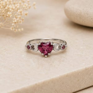 Könnte beinhalten: Ein silberfarbener Ring mit einem zentralen herzförmigen, dunkelrosa Edelstein. Die Ringschiene weist auf beiden Seiten des Herzens zwei halbmondförmige Aussparungen auf, flankiert von zwei kleineren, runden, dunkelrosa Edelsteinen.