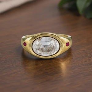 Puede incluir: Un anillo de oro con una gran piedra preciosa transparente de corte ovalado en el centro. La banda está decorada con dos pequeñas piedras preciosas redondas de color rojo vibrante, situadas a cada lado de la piedra ovalada central.