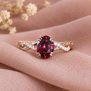 Pu&ograve; includere: Un anello in oro rosa con una gemma rosso scuro dal taglio ovale fissata da quattro griffe. La fascia presenta un design intrecciato con piccole pietre chiare incastonate a pav&eacute; lungo i lati, creando un aspetto delicato e raffinato.