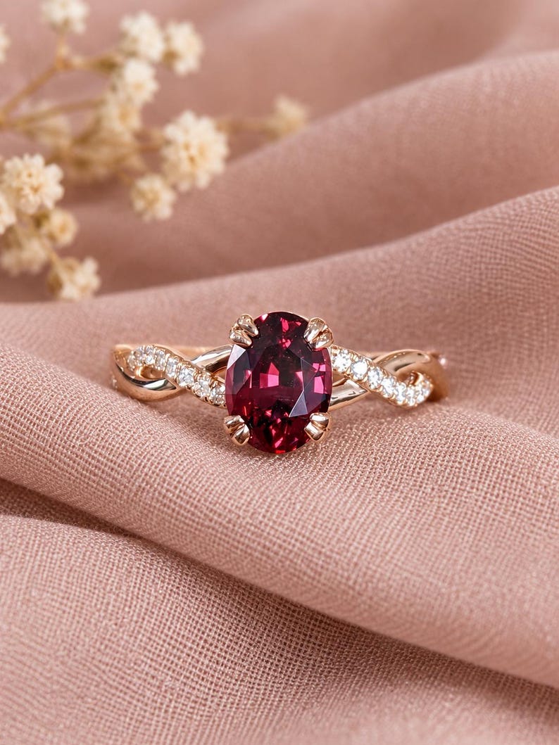 Pu&ograve; includere: Un anello in oro rosa con una gemma rosso scuro dal taglio ovale fissata da quattro griffe. La fascia presenta un design intrecciato con piccole pietre chiare incastonate a pav&eacute; lungo i lati, creando un aspetto delicato e raffinato.