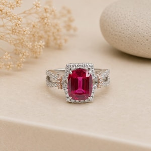Pode incluir: Um anel de prata com uma grande pedra preciosa retangular rosa-escura no centro, rodeada por um halo de pequenas pedras brancas. A banda é dividida em duas secções, acentuada com detalhes em ouro rosa e incrustada com pequenas pedras brancas.