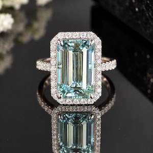 May include: A ring featuring a large, rectangular, emerald-cut light blue gemstone set in a halo of small, round, clear stones. The band is also encrusted with small, round, clear stones, and the entire piece is crafted from a metallic material.