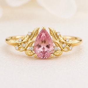 Könnte beinhalten: Ein goldener Ring mit einem zentralen birnenförmigen rosa Edelstein. Die Ringschiene ist mit metallischen Blattmotiven gestaltet, die vom Zentrum ausgehen und mit kleinen, runden, klaren Steinen entlang des goldenen Laubs besetzt sind.
