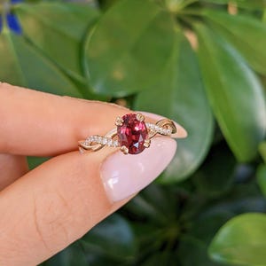 Pu&ograve; includere: Un anello in oro con una pietra preziosa rosso scuro dal taglio ovale al centro, fissata da quattro griffe. La fascia presenta un design intrecciato con piccole pietre chiare incastonate a pav&eacute; sui lati, creando un effetto scintillante.