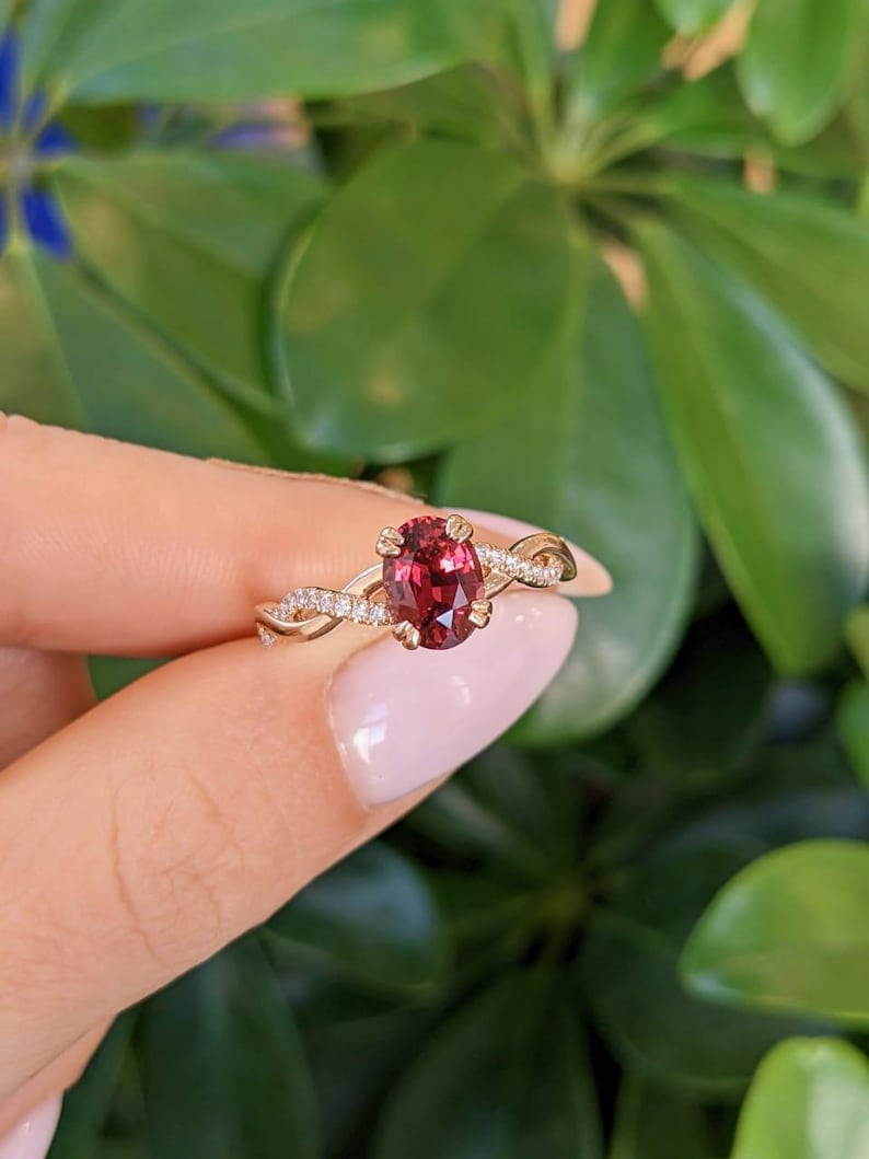 Pu&ograve; includere: Un anello in oro con una pietra preziosa rosso scuro dal taglio ovale al centro, fissata da quattro griffe. La fascia presenta un design intrecciato con piccole pietre chiare incastonate a pav&eacute; sui lati, creando un effetto scintillante.