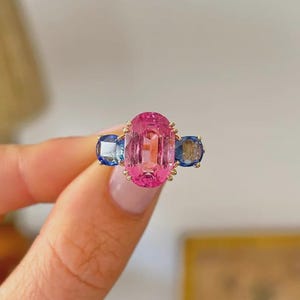 Puede incluir: Un anillo de tres piedras con una gema central grande de color rosa y corte ovalado, flanqueada por dos gemas azules más pequeñas de corte redondo. Las piedras están montadas en una banda de metal dorado con garras delicadas.
