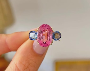 Può includere: Un anello a tre pietre con una grande gemma centrale rosa dal taglio ovale, affiancata da due gemme blu più piccole dal taglio rotondo. Le pietre sono incastonate in una fascia di metallo dorato con delicate griffe di fissaggio.