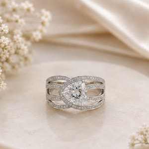 May include: A silver-coloured ring featuring a large, central pear-shaped clear stone surrounded by multiple bands encrusted with small, sparkling white pavé stones. The design incorporates overlapping, curved metal lines that frame the centre gem.