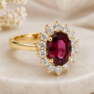 May include: A gold ring featuring a large, oval-cut, deep pinkish-red gemstone at the center, surrounded by a halo of twelve small, round, clear white diamonds set in a scalloped gold frame, with a smooth, polished gold band.