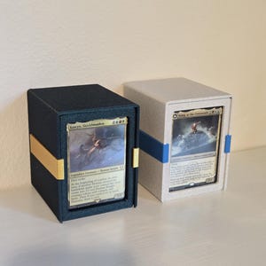 May include: Two rectangular card storage boxes with open fronts displaying trading cards. The left box is black with a gold horizontal band, holding an Eowyn, Shieldmaiden card. The right box is light gray with a blue band, holding an Aany, at the Crossroads card.