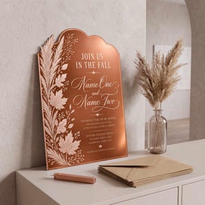 May include: An arched copper-colored wedding sign featuring white botanical illustrations of maple leaves and pampas grass. The text reads: JOIN US IN THE FALL, Name One and Name Two, SATURDAY, THE 00 OF MONTH, TWO THOUSAND TWENTY FIVE, AT FOUR O'CLOCK IN THE AFTERNOON.