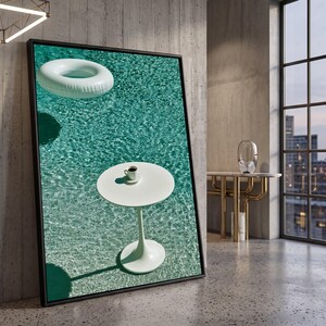 Puede incluir: Una impresión de arte mural enmarcada que muestra una mesa redonda blanca con pedestal y una pequeña taza de café encima, situada en una piscina turquesa junto a un anillo inflable blanco, con ondas de agua visibles en la superficie.