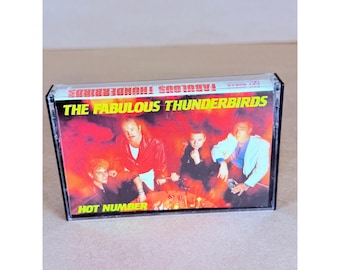 May include: A cassette tape featuring The Fabulous Thunderbirds album Hot Number. The cover art displays four band members in a red-toned setting with yellow text reading The Fabulous Thunderbirds and Hot Number at the top and bottom.