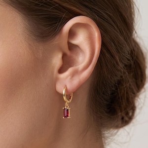 May include: A gold hoop earring featuring a small, rectangular, deep red gemstone charm suspended from the bottom. The hoop is polished gold, and the gemstone is set in a matching gold frame, creating a simple, dangling design for the earlobe.