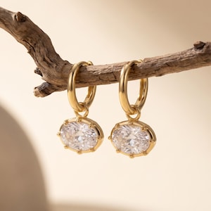 May include: A pair of gold hoop earrings, each featuring a dangling oval-shaped clear gemstone set in a gold frame. The earrings are suspended from a small, textured brown branch, showcasing a polished metallic finish and faceted stone details.