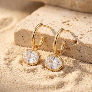 May include: A pair of gold hoop earrings featuring dangling oval-shaped clear gemstones. Each earring consists of a polished gold hoop with a prong-set, faceted oval stone suspended from the bottom, reflecting light with a brilliant white sparkle.