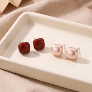 May include: Two pairs of stud earrings featuring smooth, rounded square shapes. One pair is a deep, glossy burgundy red, while the other pair is a soft, matte-finish pale peach. Each earring has a silver-toned metal post attached to the back.