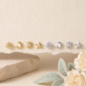 May include: A collection of eight small stud earrings arranged in a row. The set includes four gold-toned and four silver-toned pieces featuring celestial shapes: a planet with a ring, a crescent moon, a sun, and a star, each accented with clear stones.