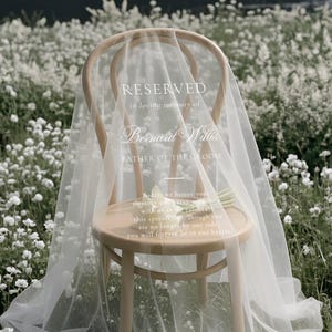 May include: A light wood chair draped with a sheer white veil featuring white printed text that reads: RESERVED in loving memory of Bernard Willis FATHER OF THE GROOM. Today we honor your memory and stay with us as we celebrate this special day.