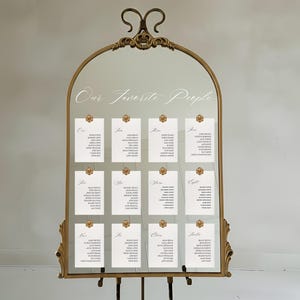 May include: An arched gold-toned metal mirror frame featuring ornate scrollwork at the top and base. The mirror displays the text Our Favorite People and twelve white rectangular cards arranged in three rows, each listing names under table numbers.