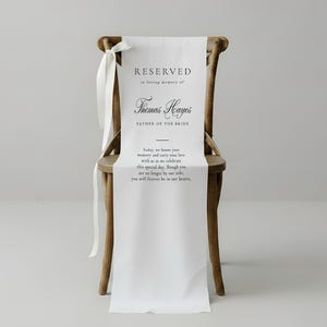 May include: A wooden chair draped with a white fabric sign featuring the text RESERVED in loving memory of Thomas Hayes FATHER OF THE BRIDE. Below the name, the text reads: Today, we honor your memory and carry your love with us as we celebrate.