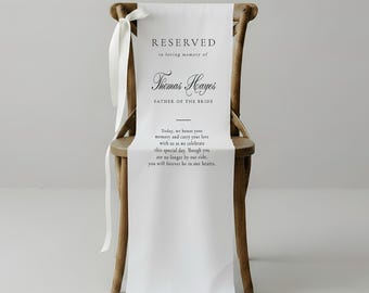 May include: A wooden chair draped with a white fabric sign featuring the text RESERVED in loving memory of Thomas Hayes FATHER OF THE BRIDE. Below the name, the text reads: Today, we honor your memory and carry your love with us as we celebrate.