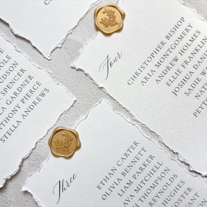 May include: White paper cards with deckled edges featuring black printed guest names and table numbers. Each card is adorned with a circular gold wax seal embossed with a botanical leaf design. The text includes table numbers Four and Three.