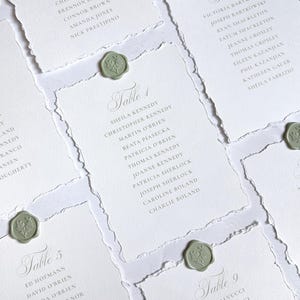 May include: A collection of white rectangular table seating cards with deckled edges, each featuring grey printed text listing guest names under specific table numbers and secured with a small, round, sage green wax seal with a floral design.
