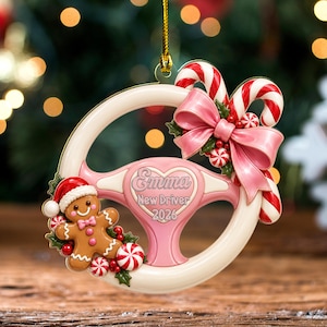 May include: A cream-colored steering wheel ornament featuring a pink heart center with the text Emma New Driver 2026. It is decorated with a gingerbread person in a Santa hat, a large pink bow, and red and white candy canes.
