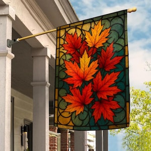 May include: A rectangular garden flag featuring a stained glass style design of vibrant orange, red, and yellow maple leaves against a dark teal and gold background. The flag is mounted on a gold metal pole attached to a white porch pillar.
