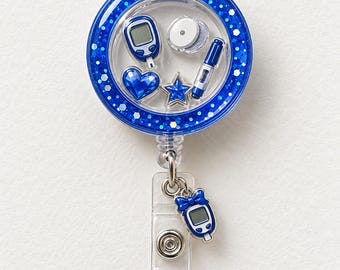 May include: A circular blue glitter badge reel featuring a clear front filled with miniature charms including a blood glucose meter, a sensor, a lancing device, a blue heart, and a blue star. A small blue glucose meter charm hangs from the clip.